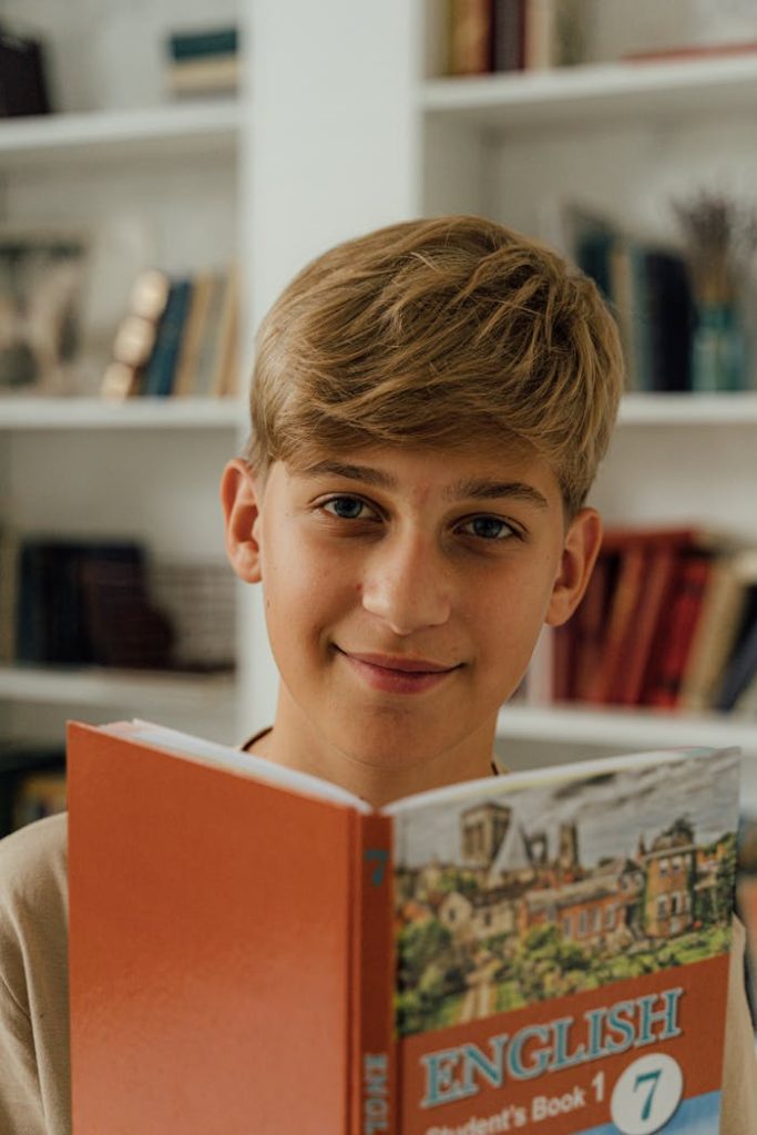 Young boy smiling while reading an English textbook indoors in a library setting.